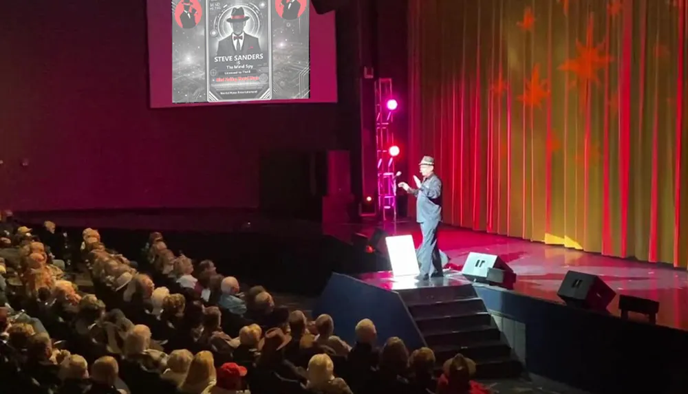 A person is performing on a stage in front of an audience with a large screen displaying promotional material in the background