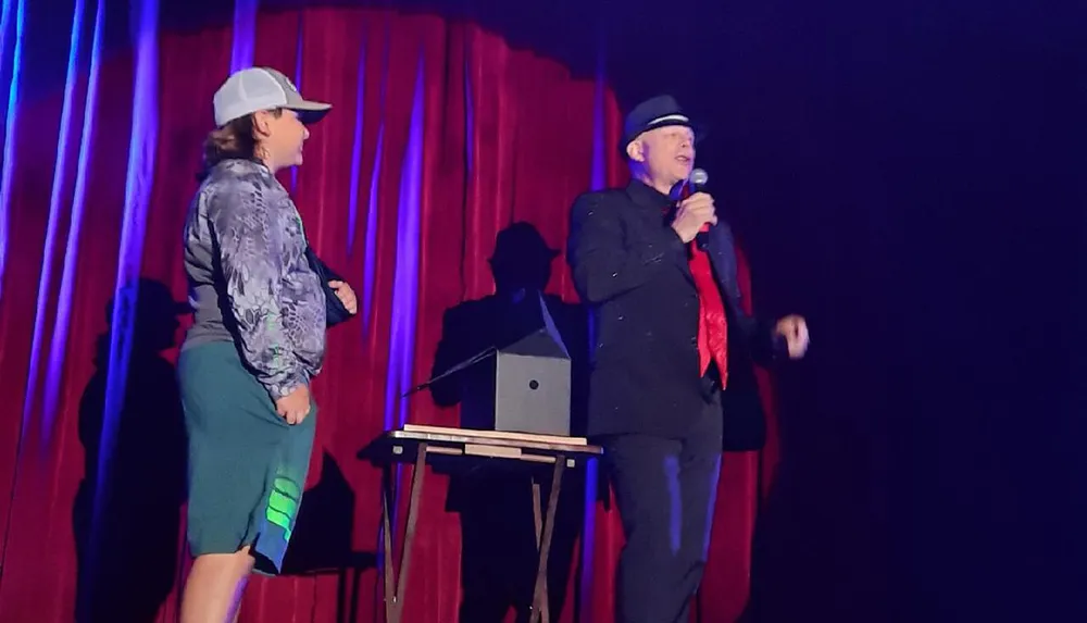 Two performers are on stage with one holding a microphone in front of red curtains