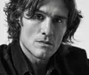 The image is a black and white close-up portrait of a person with wavy hair and a serious expression wearing a dark jacket