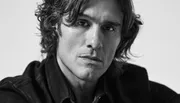 The image is a black and white close-up portrait of a person with wavy hair and a serious expression, wearing a dark jacket.