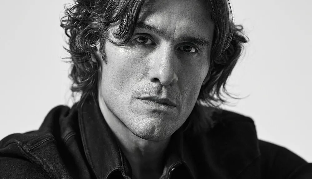 The image is a black and white close-up portrait of a person with wavy hair and a serious expression wearing a dark jacket