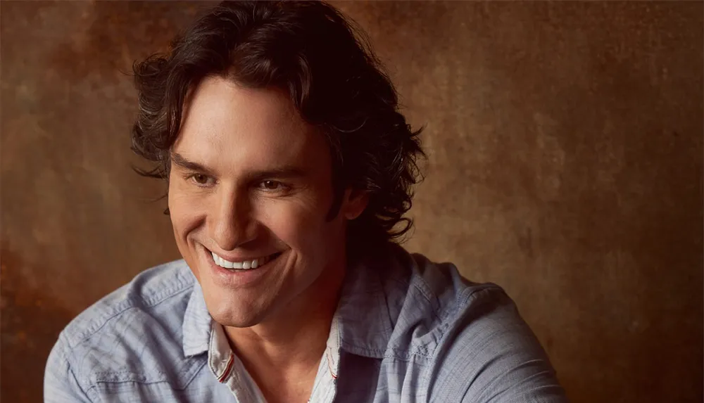 A person with curly hair is smiling and looking slightly to the side against a brown textured background