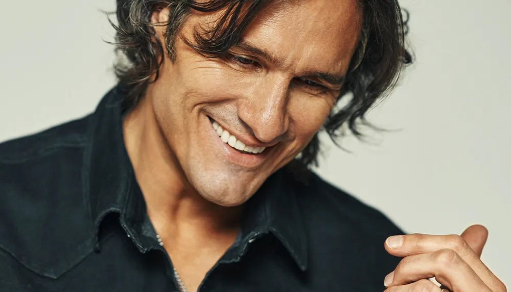 A person with long hair and a warm smile is looking downwards wearing a black shirt