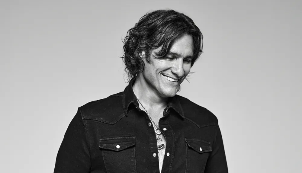 A man with wavy hair smiles while looking down wearing a button-up shirt in a black-and-white photograph