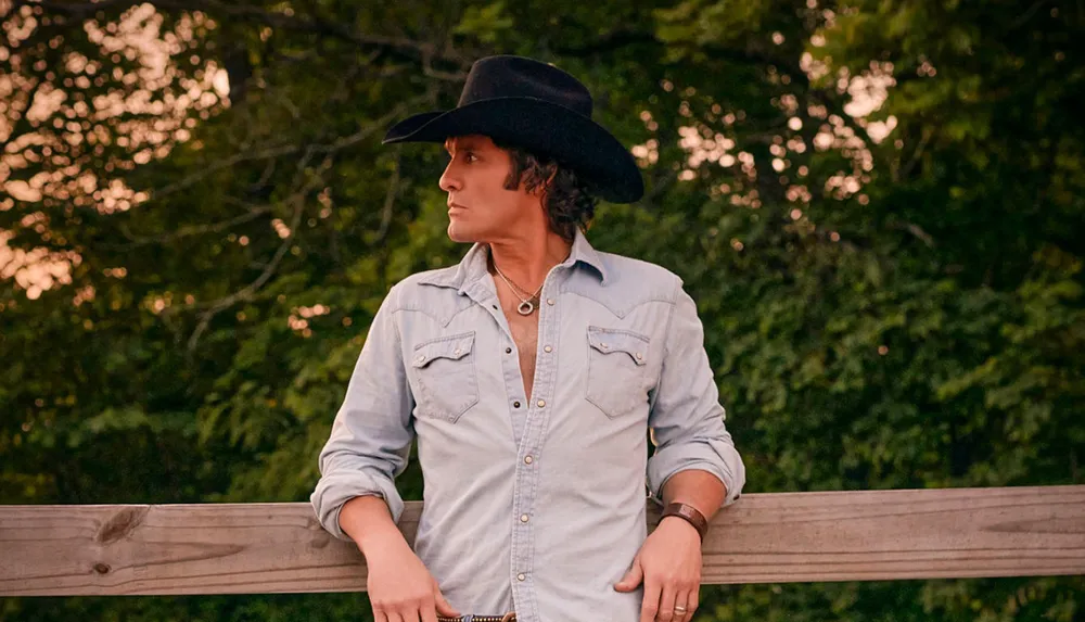 A person wearing a cowboy hat and denim shirt leans on a wooden fence looking to the side with trees in the background