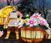 The image shows a glowing lantern sculpture of a farmer in front of a covered wagon surrounded by large illuminated flowers and barrels set against a dark wooded background