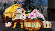 The image shows a glowing lantern sculpture of a farmer in front of a covered wagon, surrounded by large illuminated flowers and barrels, set against a dark, wooded background.