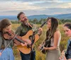 A group of four people are playing musical instruments and singing outdoors in a scenic field with mountains in the background