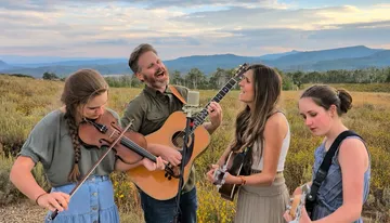 A group of four people are playing musical instruments and singing outdoors in a scenic field with mountains in the background.