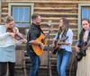 A group of four people are playing musical instruments and singing outdoors in a scenic field with mountains in the background