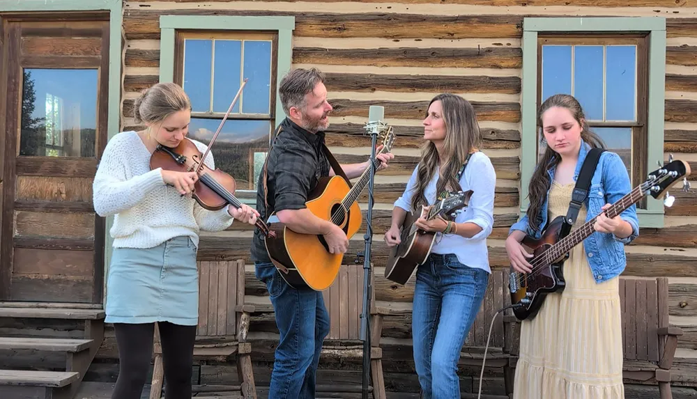 Four people are playing musical instruments outside a rustic log cabin