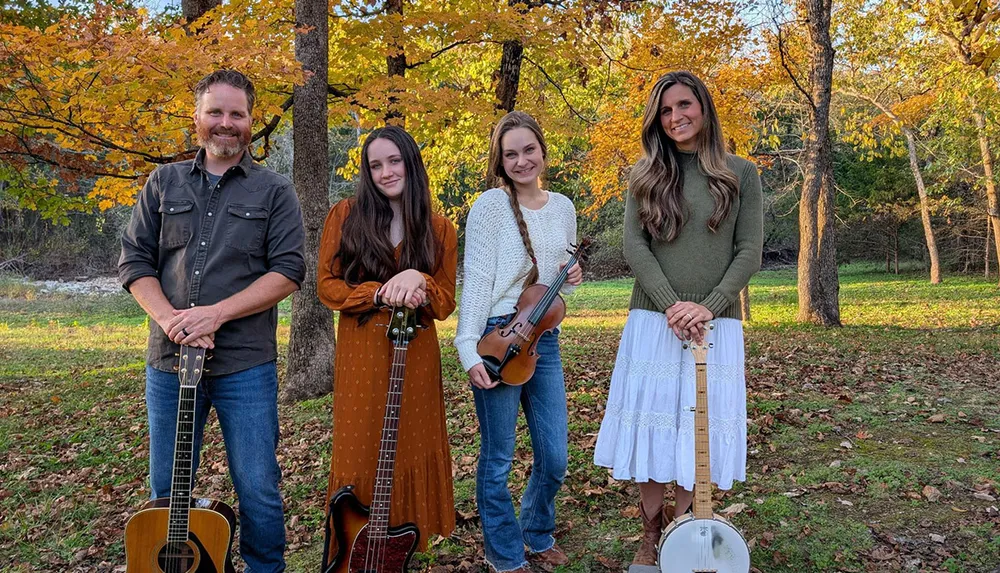Four people holding musical instruments stand together in a wooded area with autumn leaves