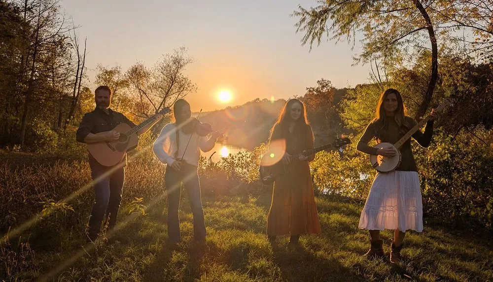 Four people are playing musical instruments in a grassy field during a sunset