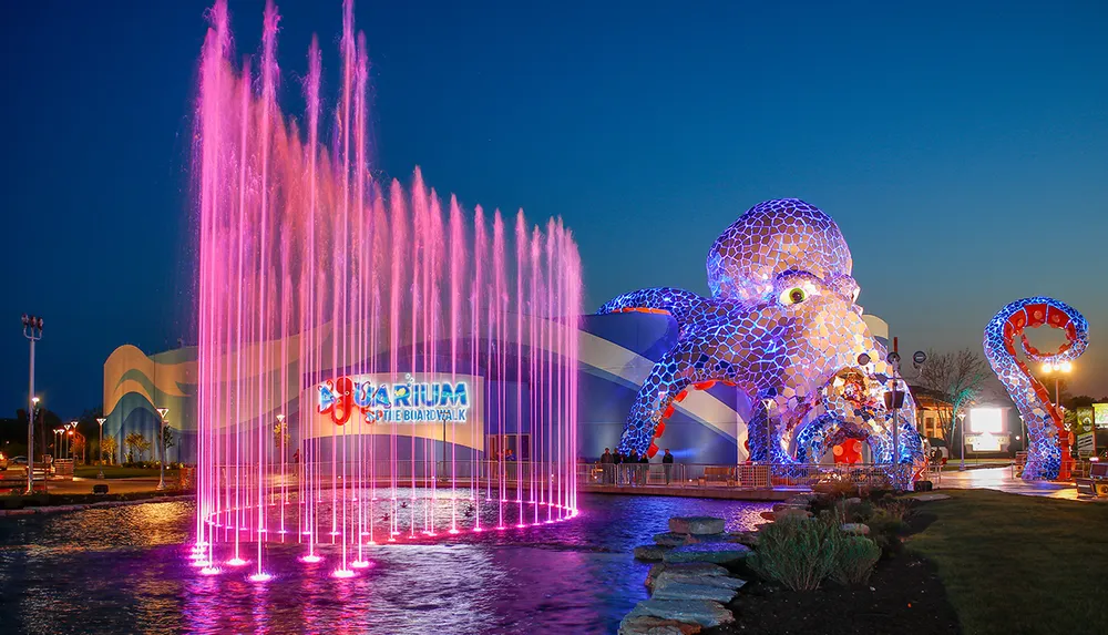A vibrant night scene features a colorful fountain display in front of an aquarium entrance adorned with a large illuminated octopus sculpture