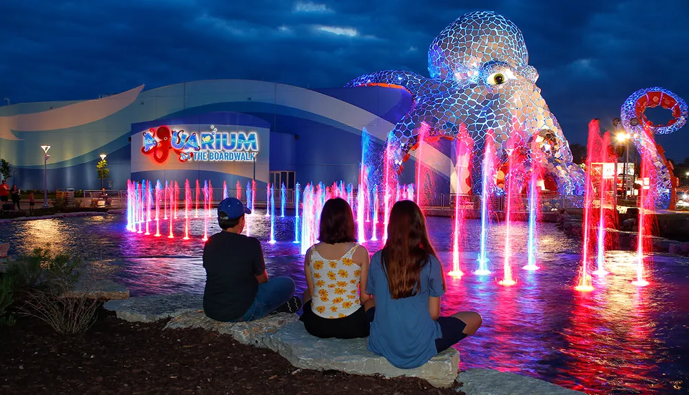 Three people sit by a brightly lit fountain in front of Aquarium at the Boardwalk featuring a large octopus sculpture