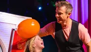 A man is playfully rubbing an orange balloon on a laughing girl's head, likely creating static electricity.
