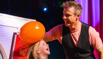 A man is playfully rubbing an orange balloon on a laughing girl's head, likely creating static electricity.