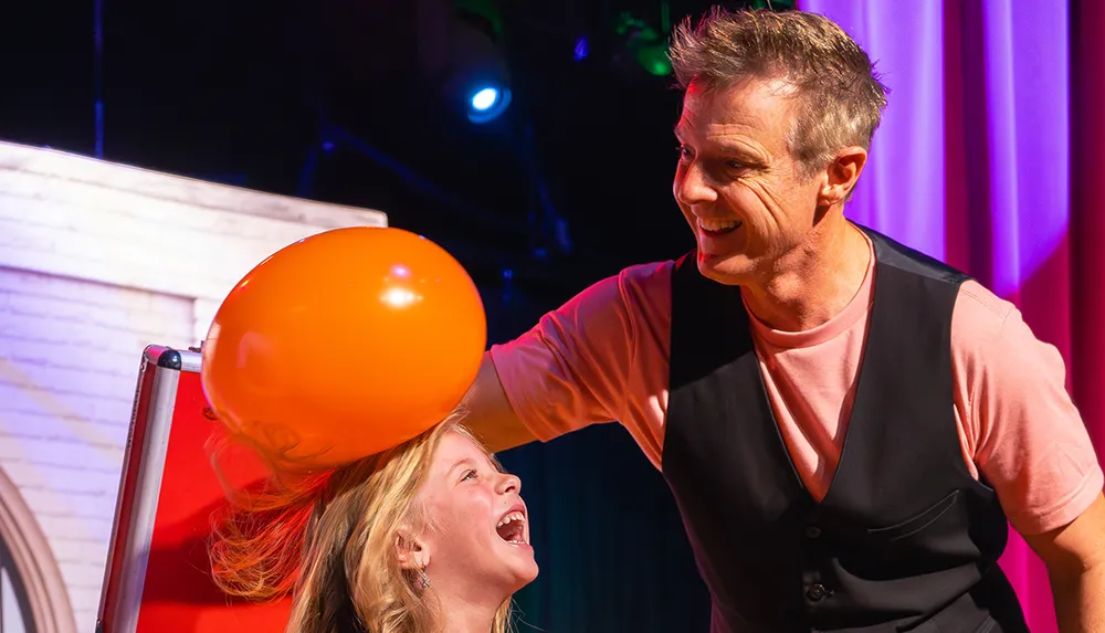 A man is playfully rubbing an orange balloon on a laughing girls head likely creating static electricity