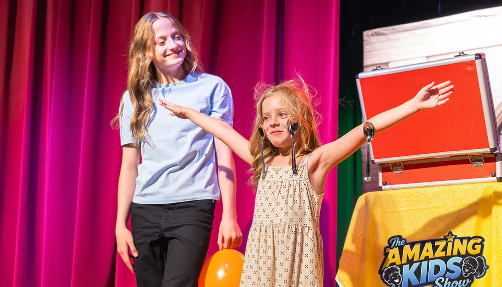 Two children are on a stage with one child smiling and the other excitedly holding out their arms with spoons on their face in front of a colorful curtain and a sign reading The Amazing Kids Show