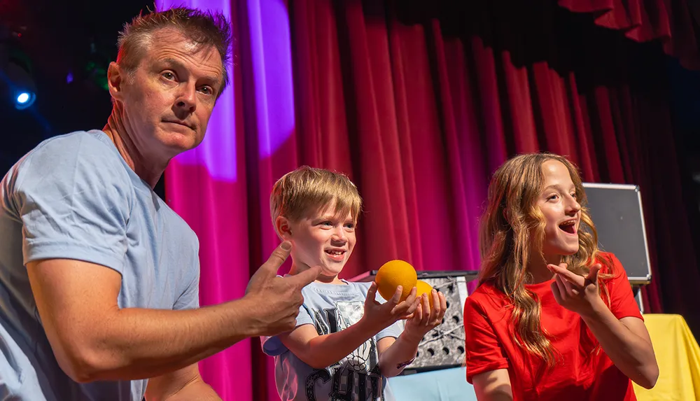 A group of three people two adults and a child are performing on stage with enthusiasm involved in a playful or theatrical activity with a small orange ball