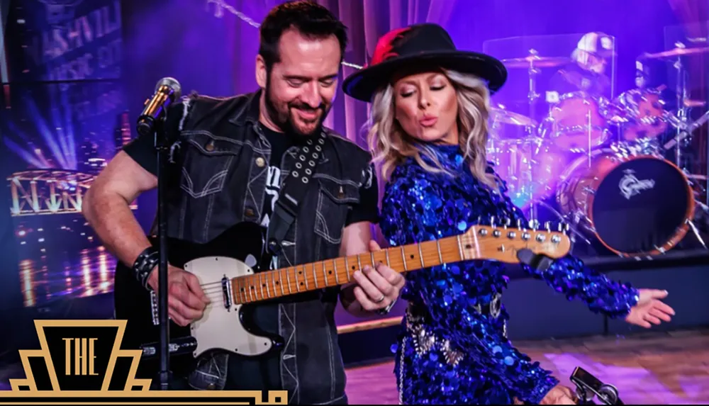 A man and a woman are energetically playing guitars on stage with a drum set and vibrant lighting surrounding them