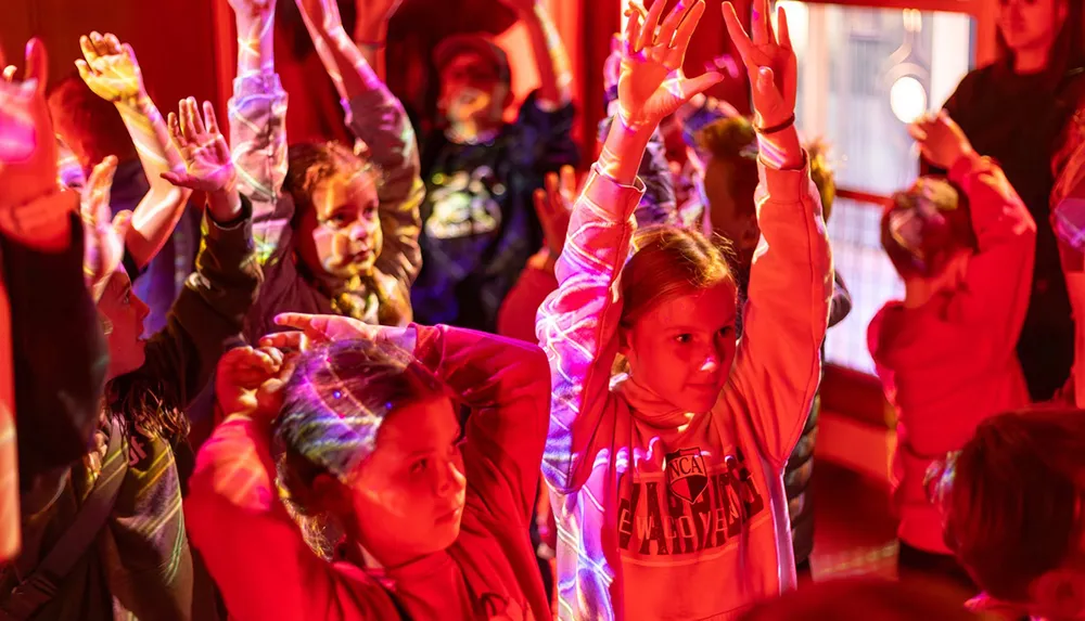 A group of children raising their arms enthusiastically in a dimly lit room with colorful projected lights