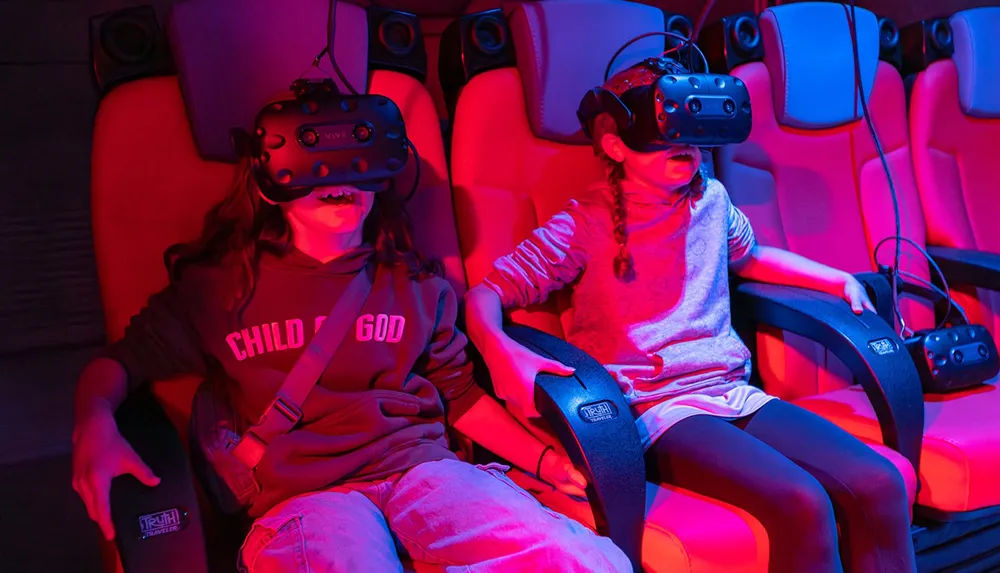 Two children wearing virtual reality headsets sit in red theater chairs appearing excited and engaged in a VR experience