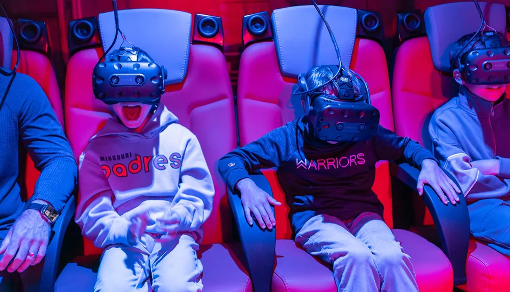 Several people are seated in red-lit chairs wearing virtual reality headsets appearing engaged and animated