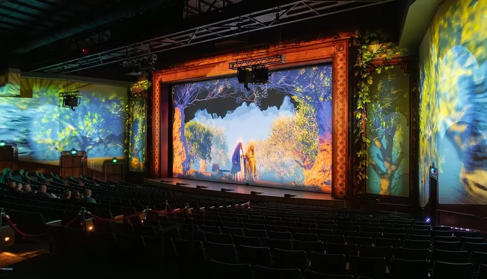 The image shows a theater with a vibrant stage and projected visuals of a colorful fantastical forest scene surrounded by an empty audience area