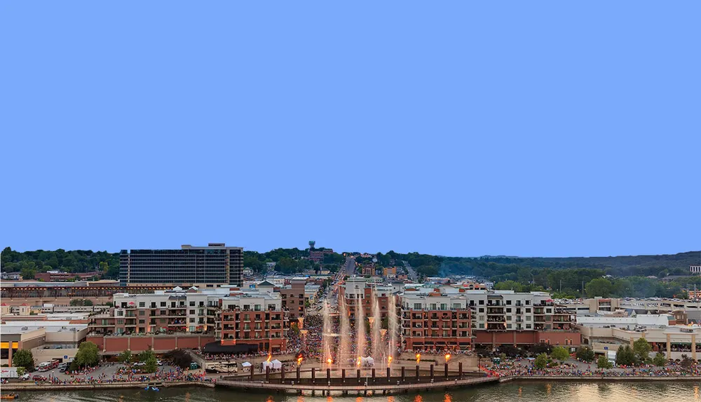 The image shows a cityscape with people gathered around a fountain shooting water high into the air against a clear blue sky