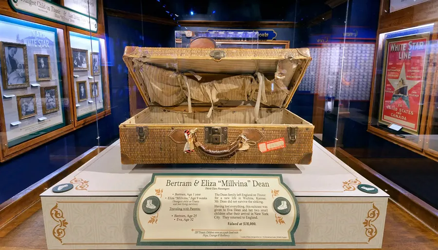 The image shows a worn, historical suitcase displayed in a museum, accompanied by informational text about Bertram and Eliza Millvina Dean, Titanic's youngest survivor.