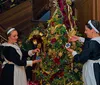 Two women in traditional maid outfits are decorating a lavishly adorned Christmas tree with ornaments near a grand staircase