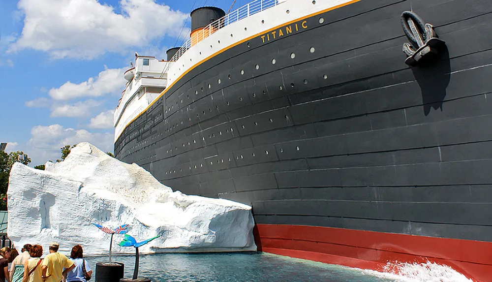 This image features a group of people looking at a large stationary replica of the Titanic ship next to an artificial iceberg likely part of a themed attraction or exhibit