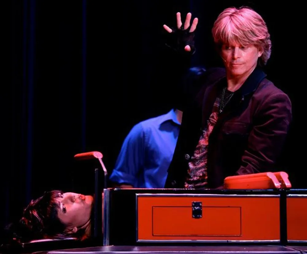 A magician is performing a classic sawing a person in half illusion on stage with a woman lying in the box and the magician gesturing dramatically with his hand raised