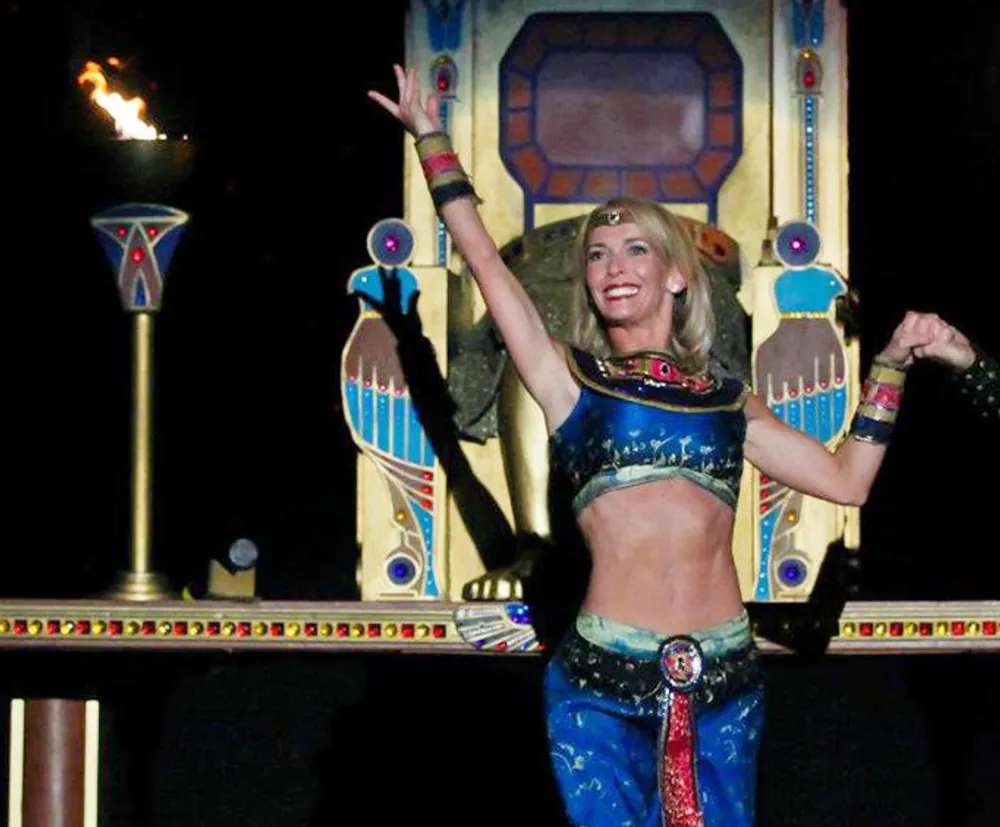 A person in Egyptian-themed costume is performing with a smile against a backdrop featuring Egyptian-style decorations and a lit torch