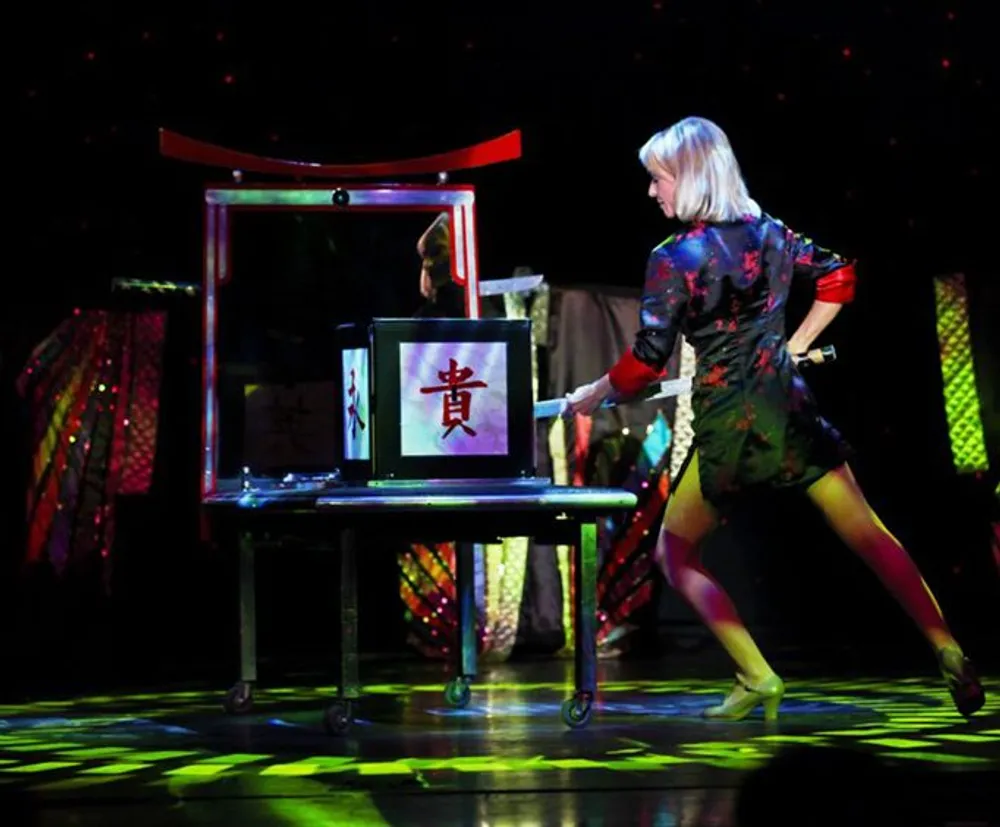 A performer is pushing a transparent box on a wheeled table with an Asian character displayed on it during what appears to be a magic show or theatrical performance