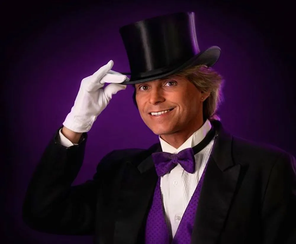 A person dressed in a formal magicians attire with a top hat bow tie and white gloves is smiling at the camera against a purple backdrop