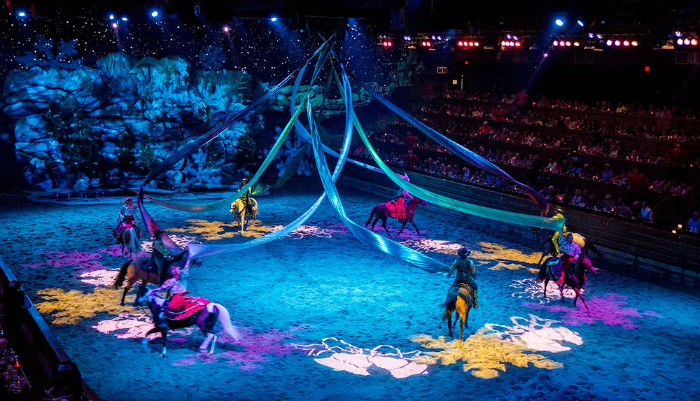 Riders on horseback perform in an equestrian-themed show before an audience in an arena illuminated with colorful lighting and decorated with theatrical scenery