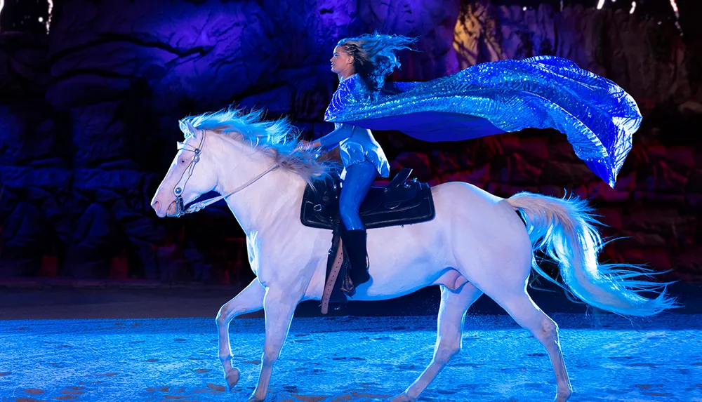 A person in a sparkling blue costume rides a white horse whose mane appears tinted blue set against a dramatic dimly lit rocky backdrop