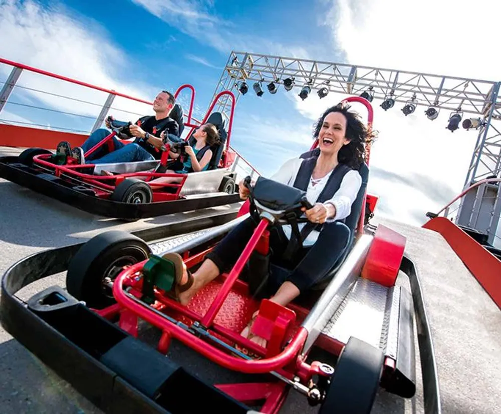 Three people are enjoying a go-kart race under a clear sky with a structure that appears to be part of a race track or entertainment facility in the background