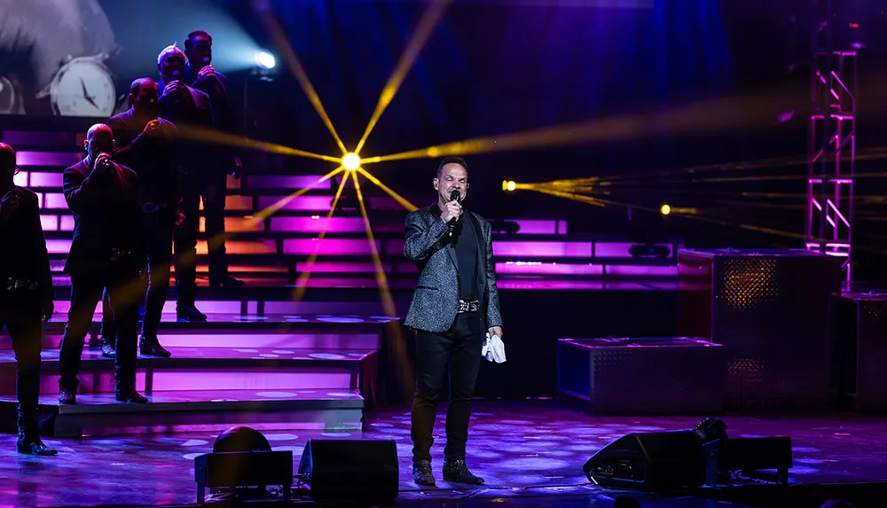 A performer sings on stage with a microphone in hand while a band appears illuminated by colorful stage lighting in the background