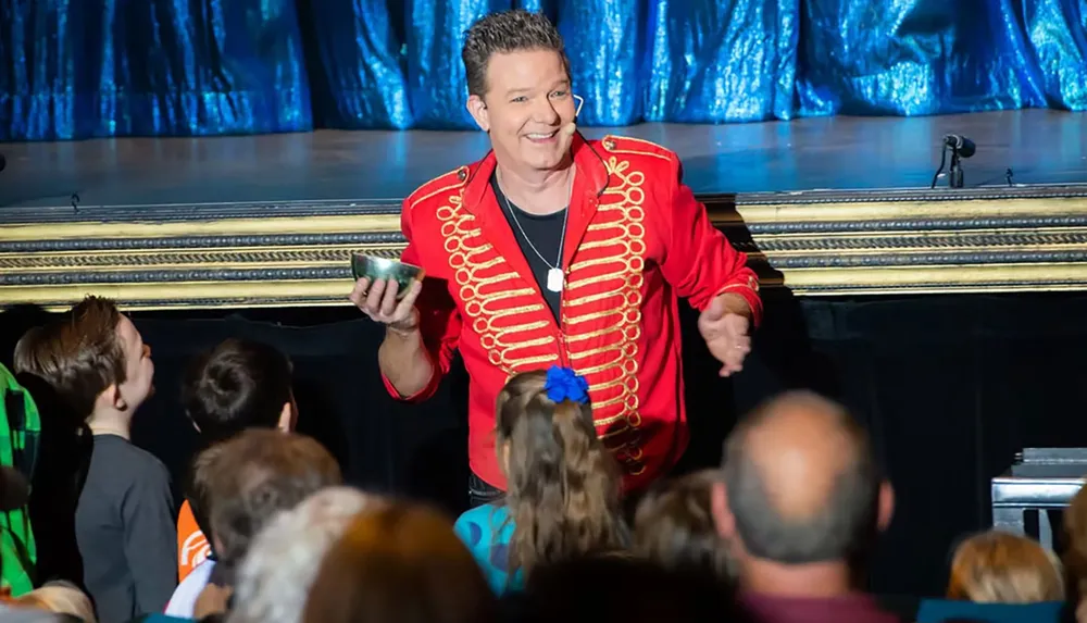 A performer in a red jacket is smiling and engaging with an audience from the stage