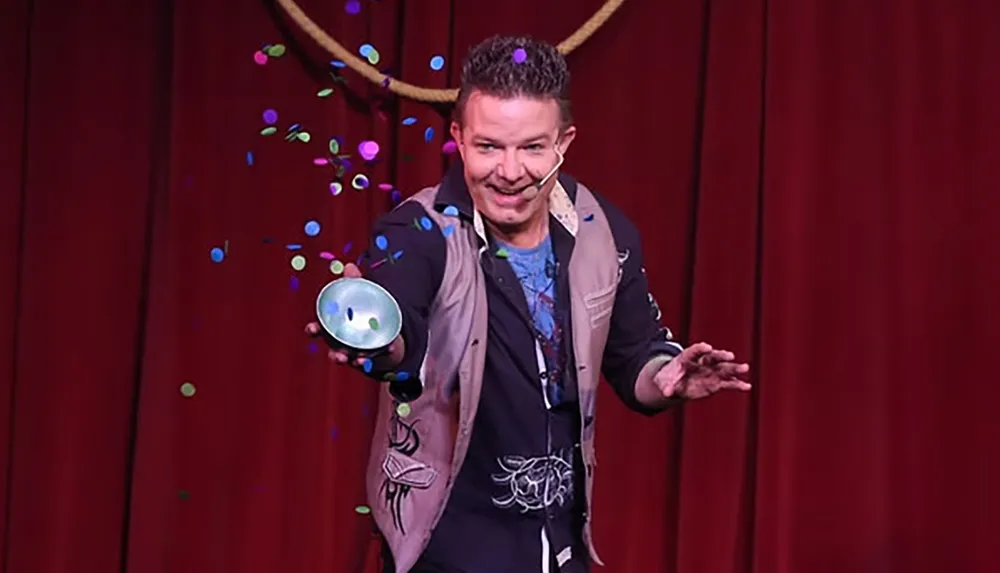 A performer on stage is playfully scattering colorful confetti from a small device set against a red curtain backdrop