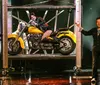 A performer in a bright costume dramatically presents a yellow motorcycle with a person lounging on it surrounded by other performers on stage