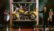 A performer in a bright costume dramatically presents a yellow motorcycle with a person lounging on it, surrounded by other performers on stage.
