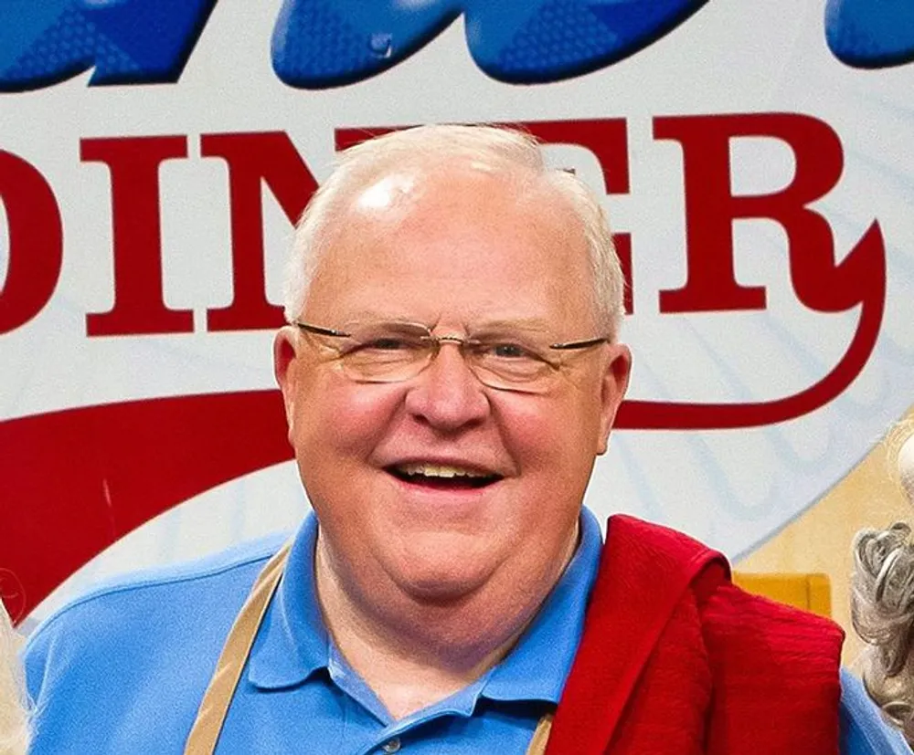 The image shows a cheerful balding man with glasses wearing a blue shirt and holding a red cardigan over his left shoulder with a branded background partially visible behind him