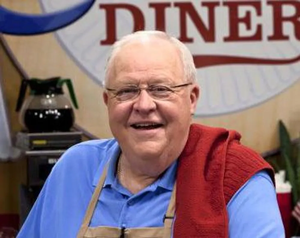 A smiling man with glasses wearing a blue shirt apron and red vest is standing in front of a sign that says DINER