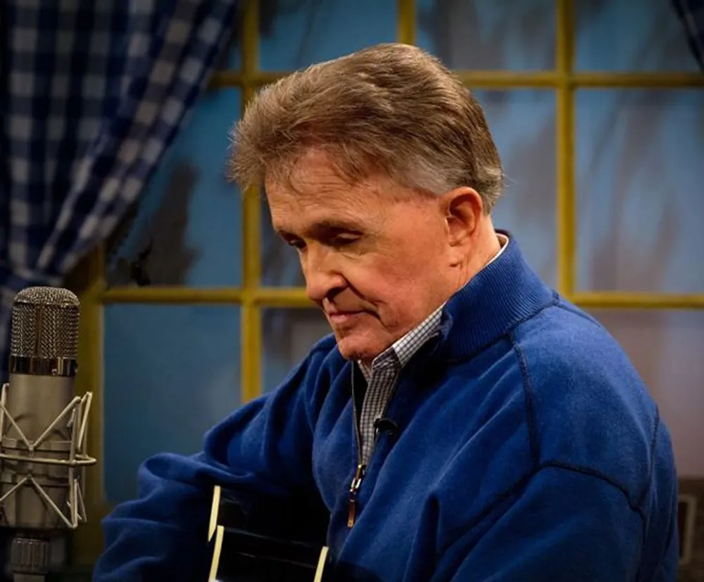 A person is focused on playing a guitar near a microphone in a room with a checkered curtain and windowpane backdrop