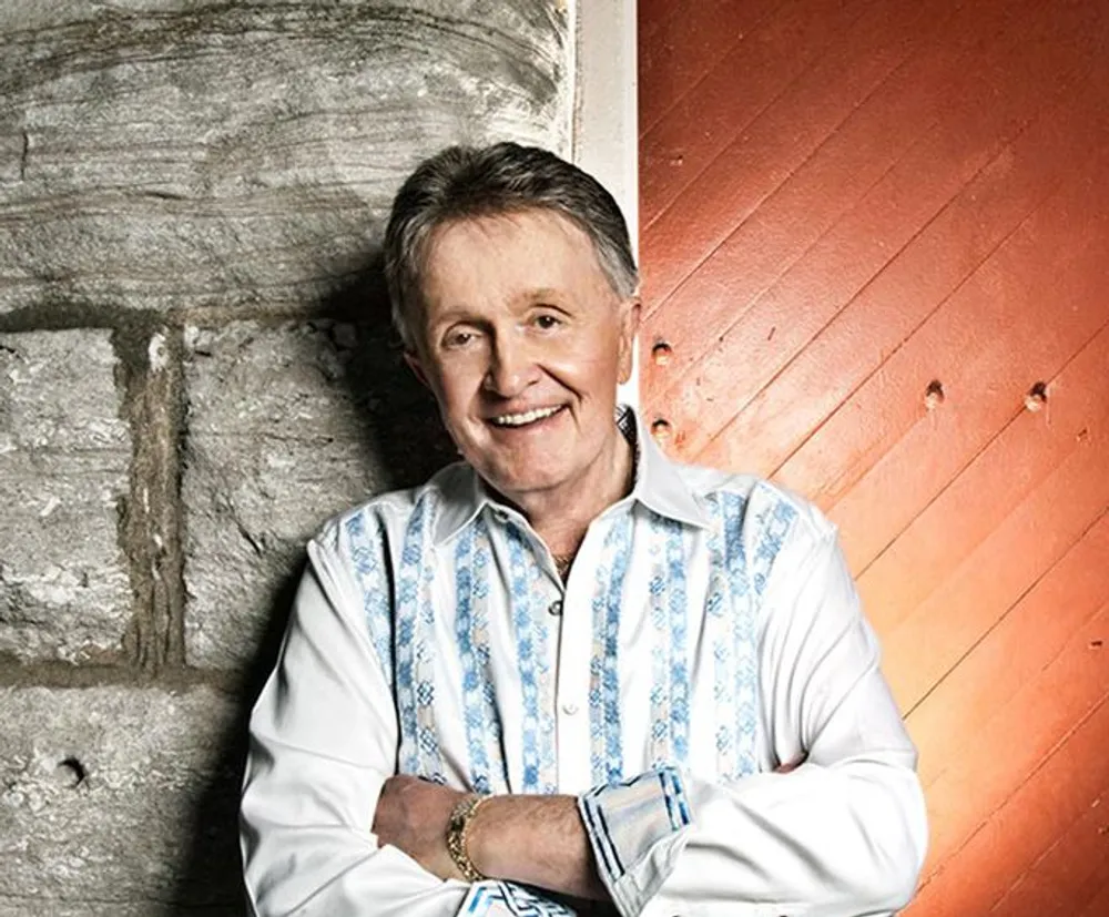 A smiling person is leaning against a rustic stone and wood wall wearing a white shirt with blue patterns and a vest exuding a casual friendly vibe