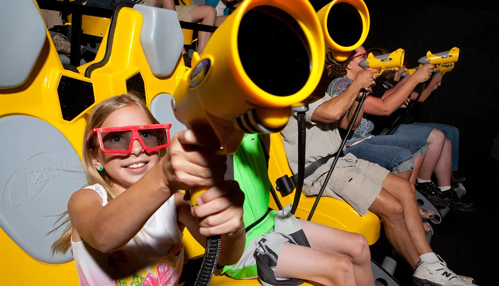 A girl wearing 3D glasses is holding a yellow toy gun while seated in a row with other people enjoying an interactive amusement ride